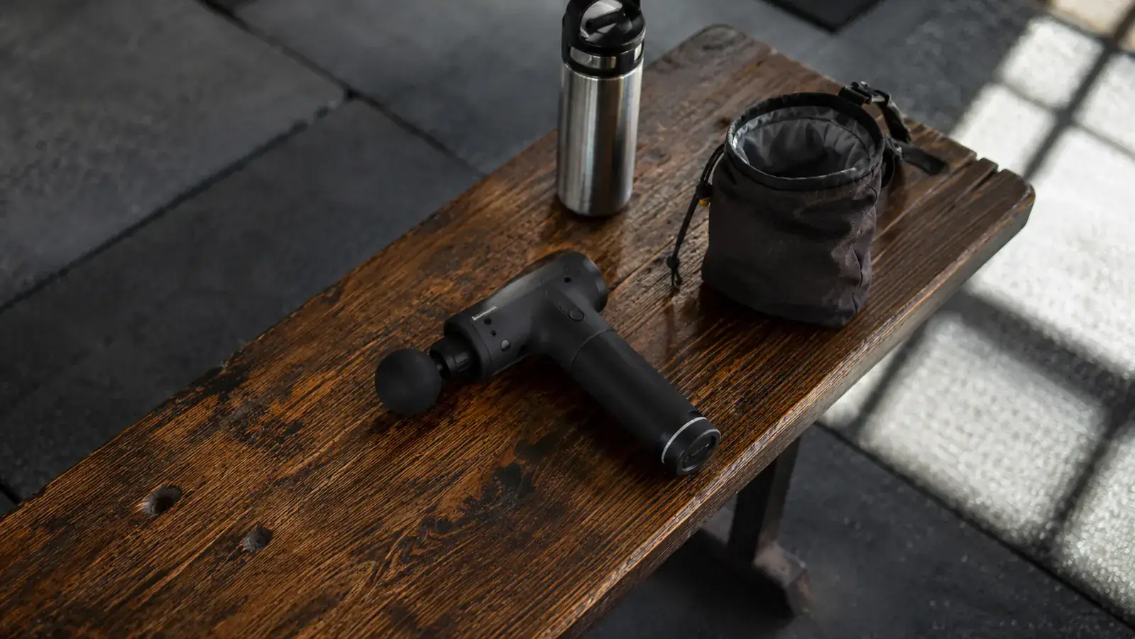 BOB AND BRAD C2 massage gun resting on a gym bench next to a water bottle and lifting chalk bag, post-workout setting