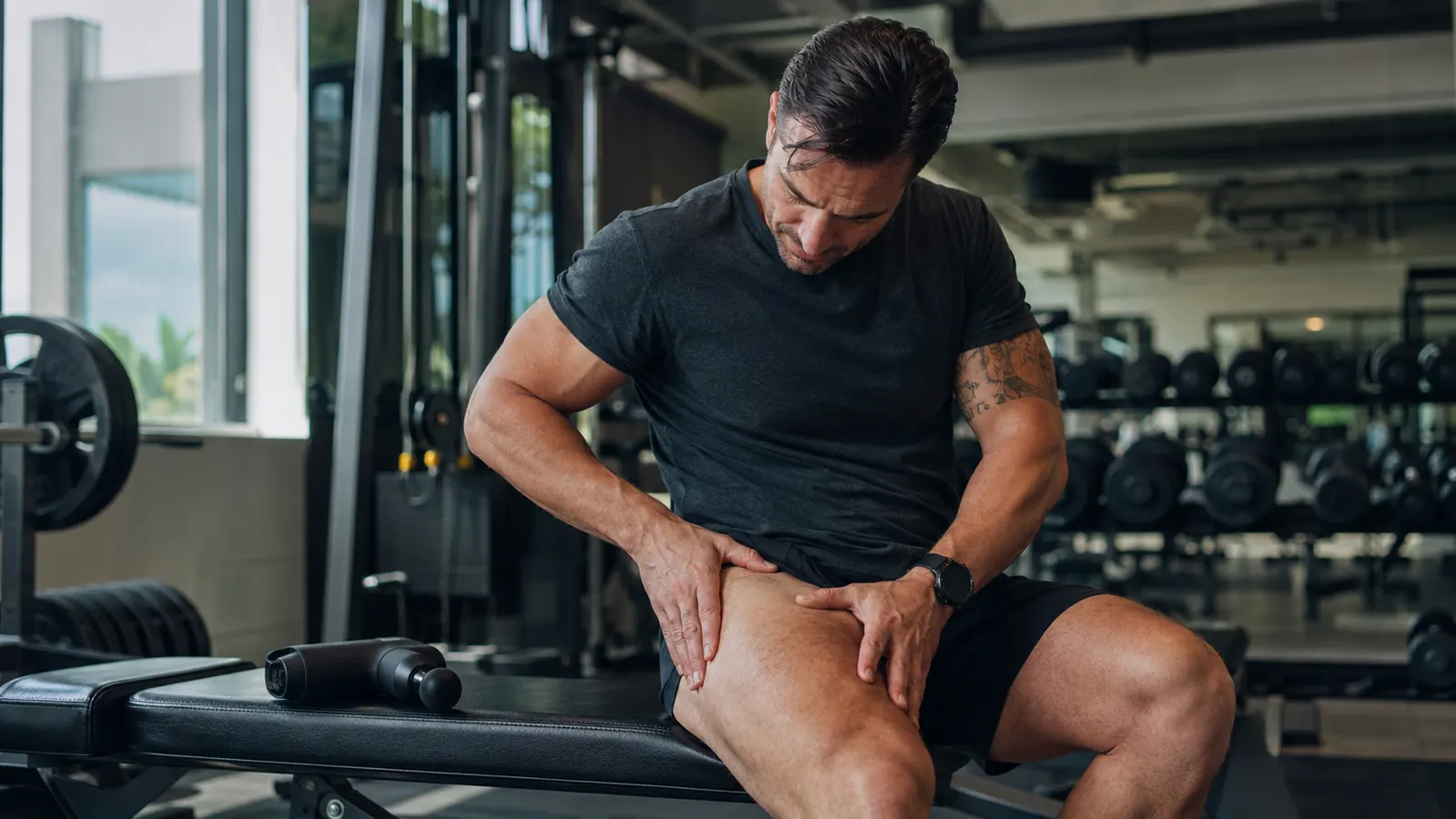 Man sitting on a gym bench resting his quad with a percussion massager nearby on the seat