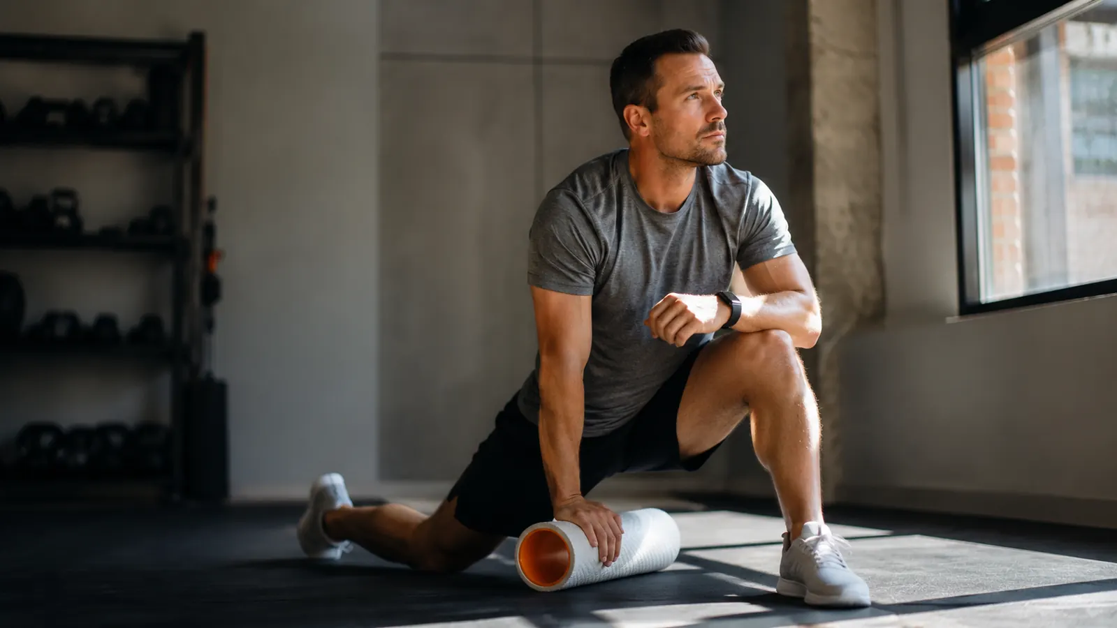 Man sitting on a gym mat in the early morning, a foam roller beside him, stretching his hip flexor before a workout