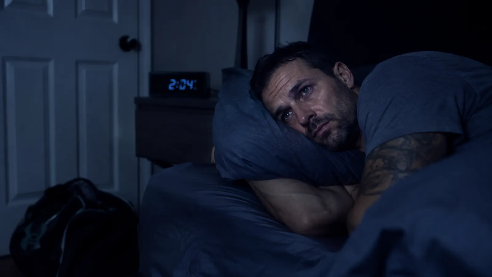 Man lying awake in bed at night, staring at ceiling, with gym bag visible on floor